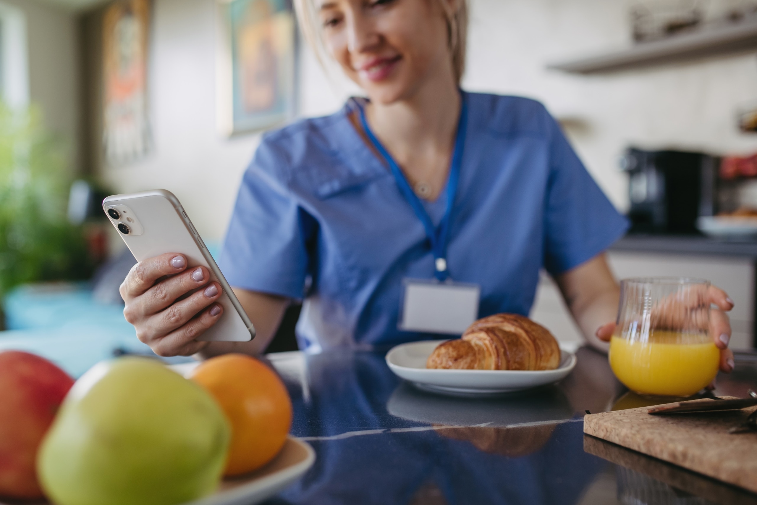 Nurse in scrubs at home using a mobile phone and eating snacks
